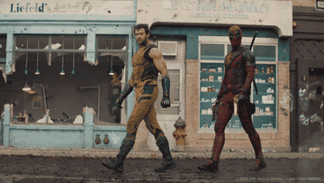 Hugh Jackman as Wolverine in a yellow suit and Ryan Reynolds as Deadpool in a red and black suit walk together in a damaged street in front of shops