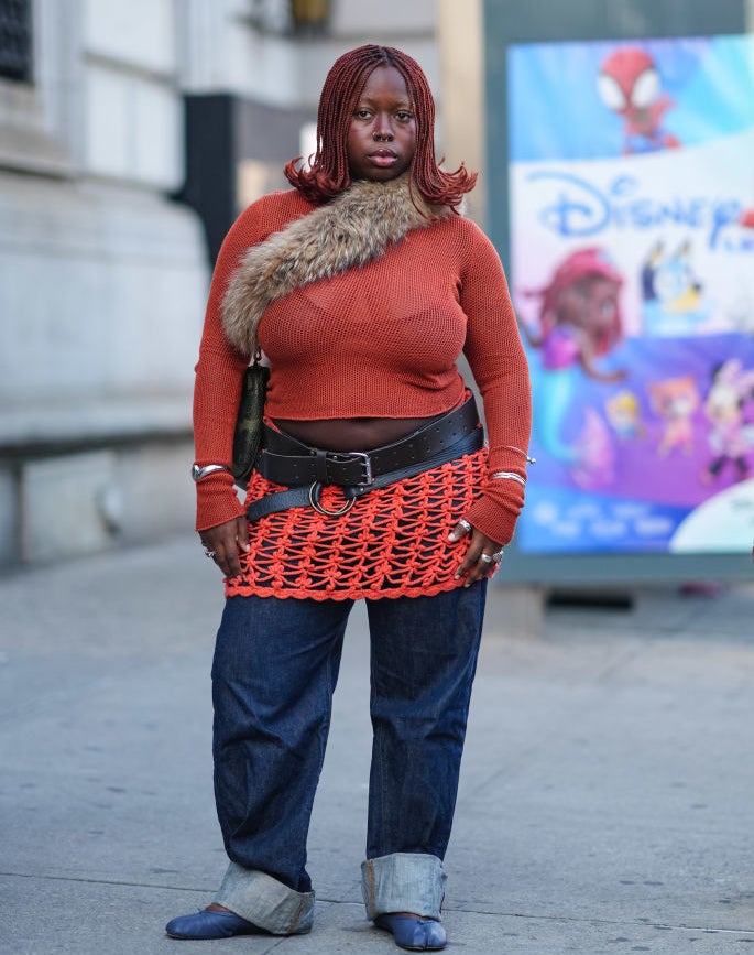 A fashion show attendee with red braided hair is wearing a cropped knit sweater with a fur detail, a belt, a red lattice skirt, and jeans