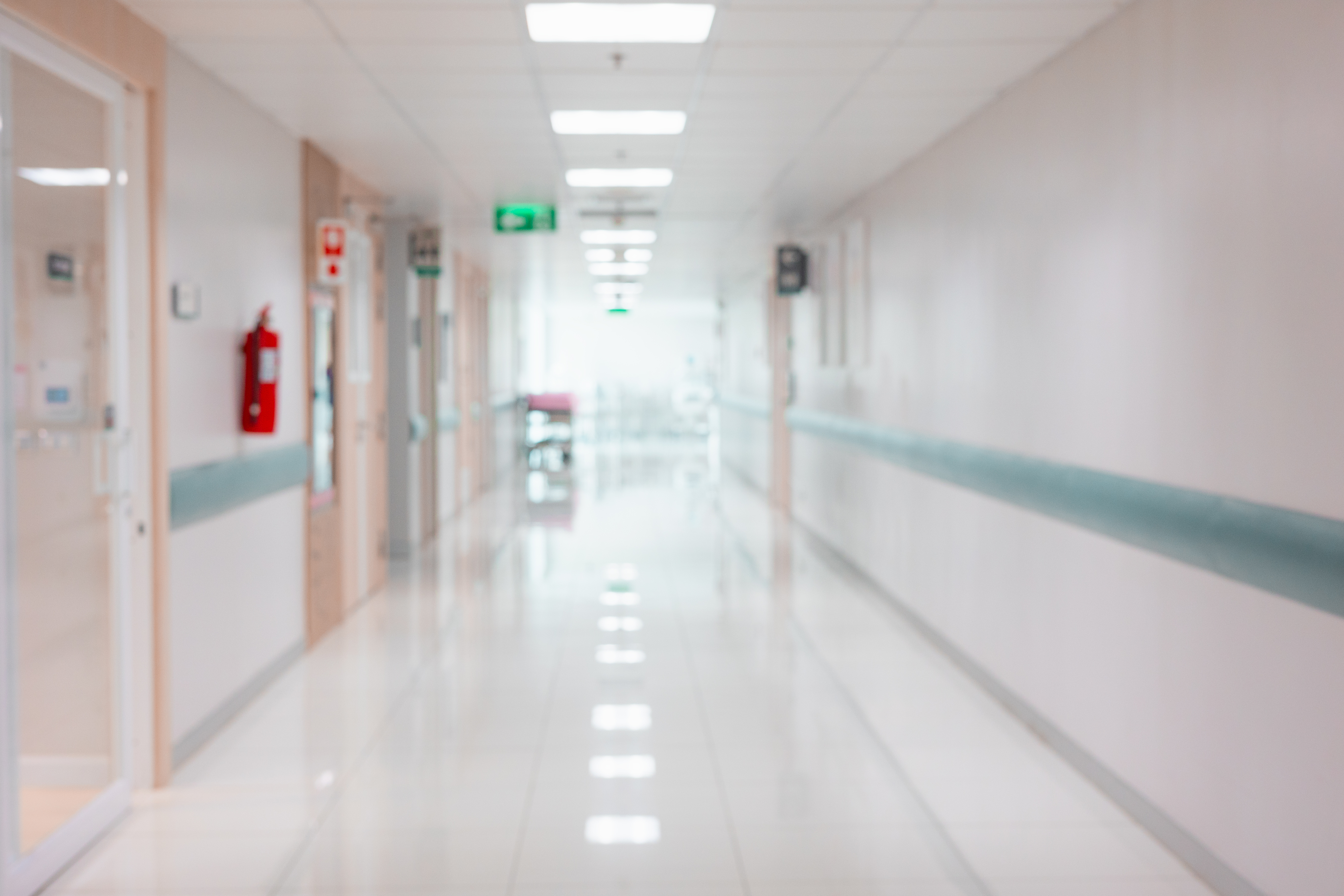 A bright hospital hallway with closed doors, a red fire extinguisher on the wall, and green exit signs visible above