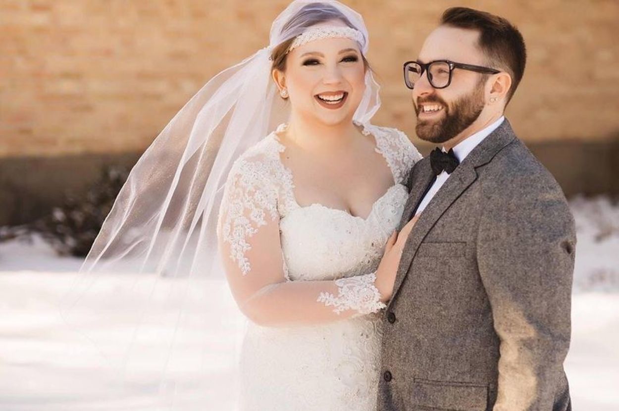 Bride in a lace wedding dress and groom in a gray suit with a bow tie smiling together outside in the snow