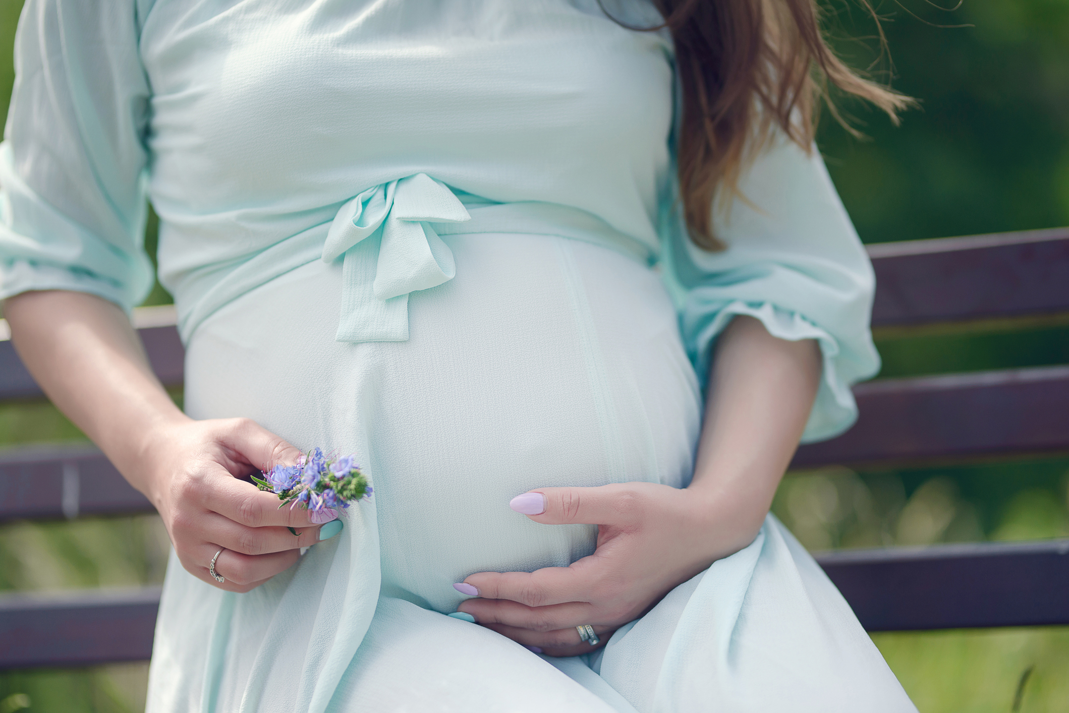 Close-up of a pregnant woman sitting on a bench, gently holding small flowers in one hand and cradling her belly with the other