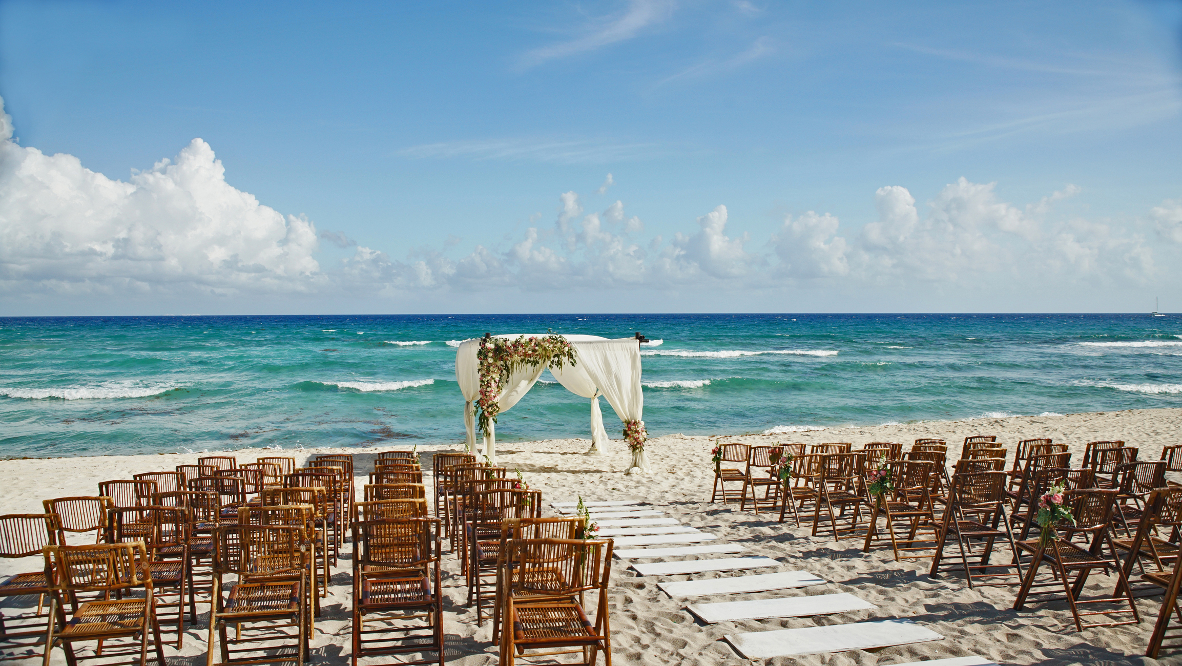 An outdoor beach wedding setup features rows of wooden chairs facing a floral-decorated arch by the ocean