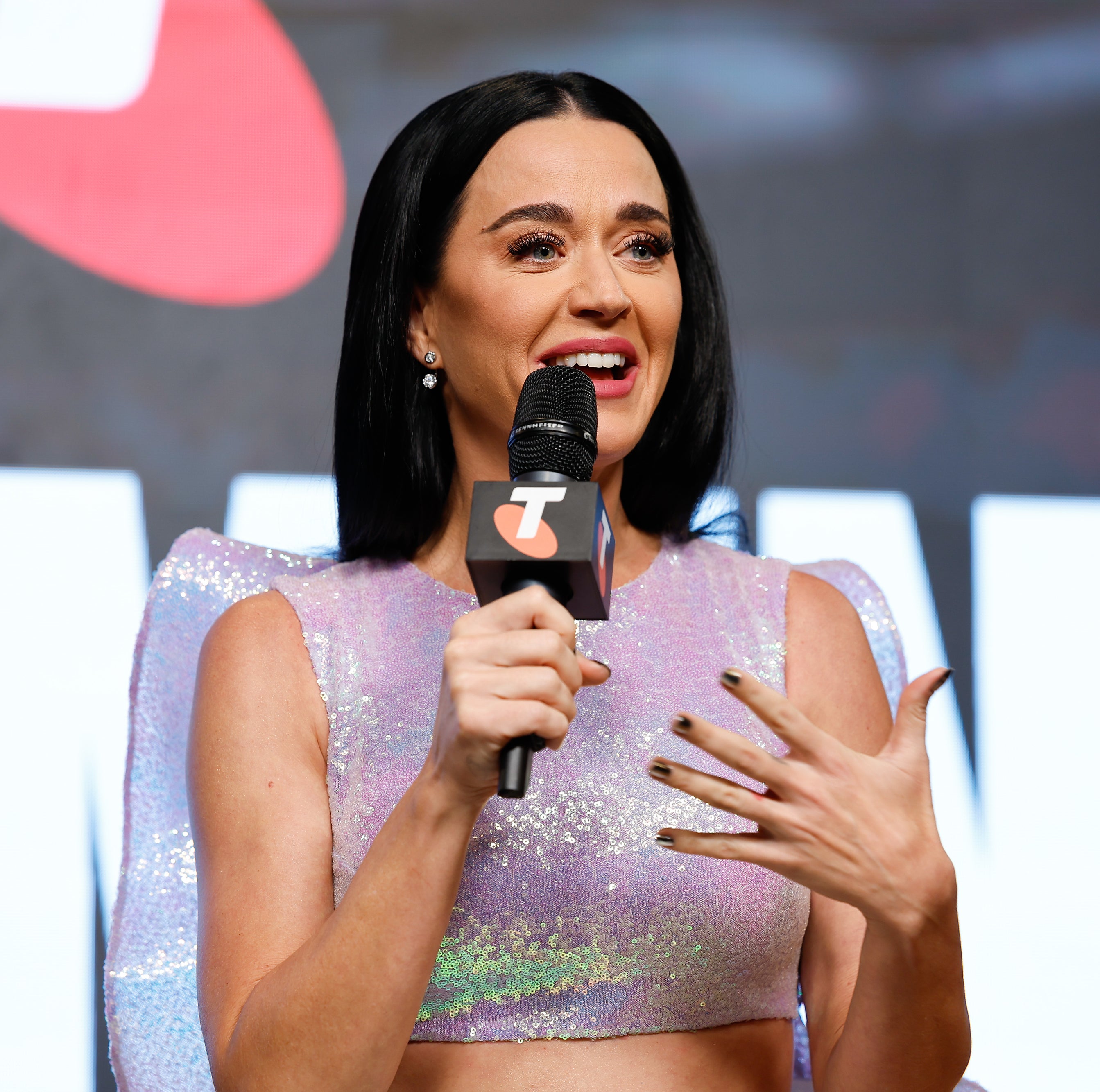 Katy Perry speaks into a microphone on stage, wearing a sparkling top with structured shoulders