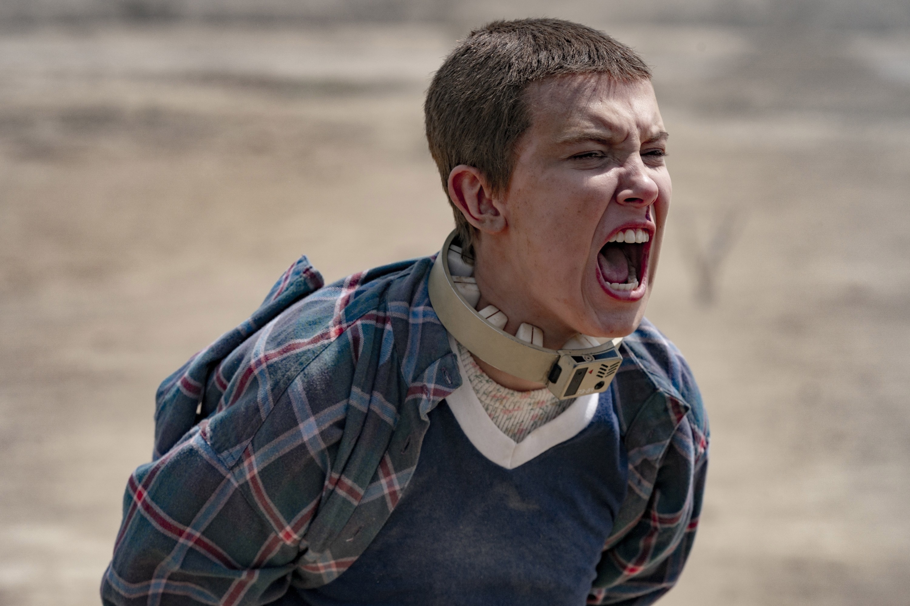 Millie Bobby Brown as Eleven in &quot;Stranger Things&quot; screams with a fierce expression, wearing a plaid shirt and a collar, in a dramatic outdoor scene