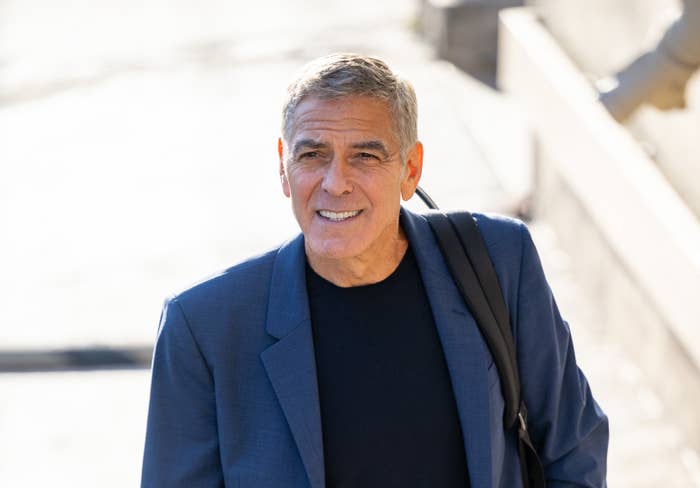 George Clooney, wearing a dark suit jacket over a black shirt, smiling while looking slightly off-camera. He has a bag slung over his shoulder