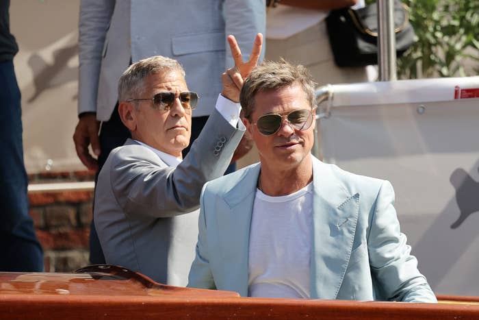 George Clooney flashes a peace sign behind Brad Pitt's head while both wear suits and sunglasses onboard a yacht