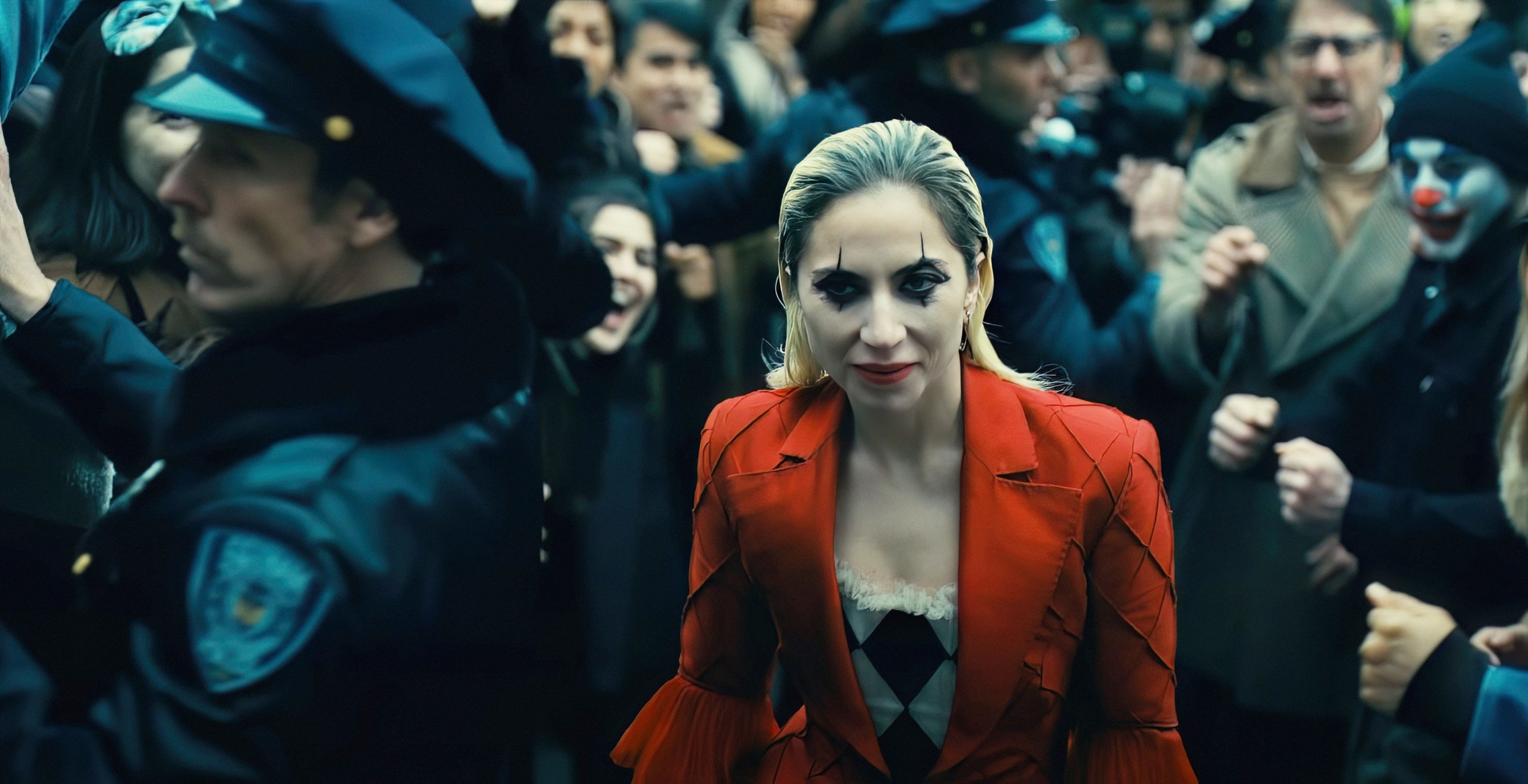 Lady Gaga, dressed in a jacket with striped accents, walks through a crowded scene filled with police officers and onlookers
