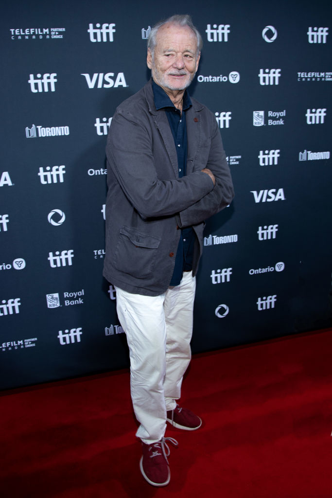 Bill Murray stands on the red carpet wearing a casual jacket, dark shirt, light pants, and red sneakers at the Toronto International Film Festival