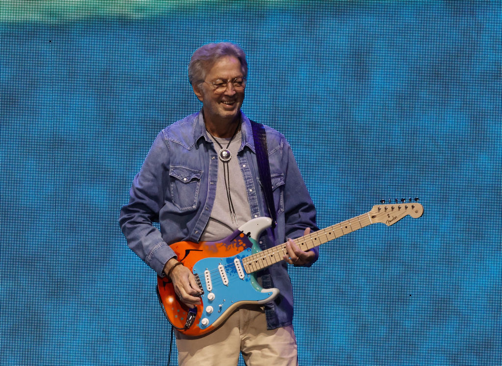 Eric Clapton plays an electric guitar on stage, wearing a casual button-up shirt and a necklace