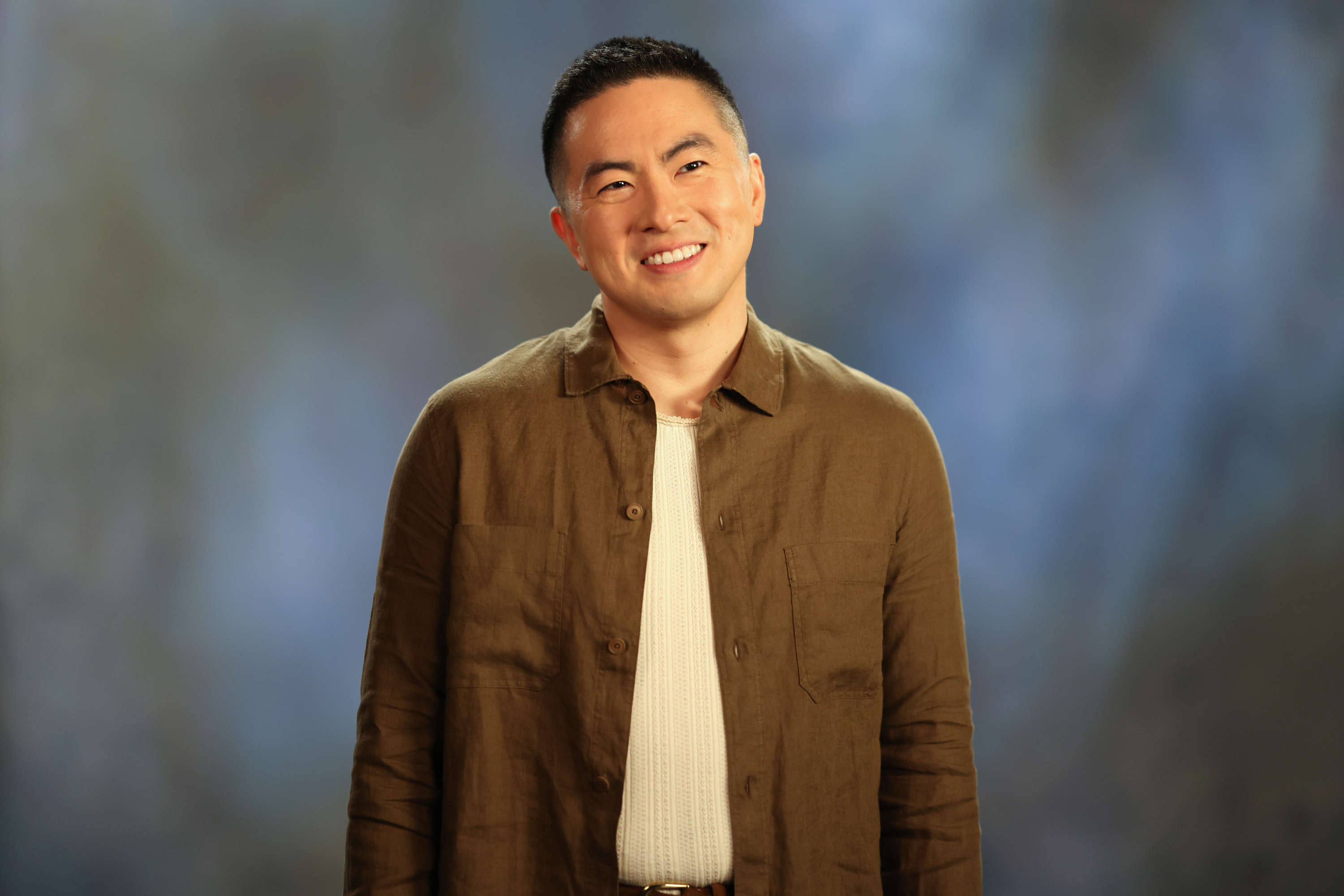 Bowen Yang is photographed smiling against a blurred background. They are dressed casually in a button-up shirt and a light, textured top