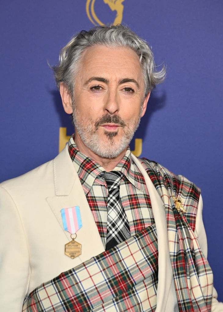 Alan Cumming on the red carpet, wearing a beige blazer with a tartan sash and tie, and a medal pinned to his lapel