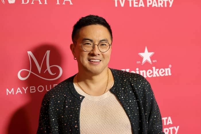 Bowen Yang at the BAFTA TV Tea Party, wearing glasses, a shirt, and a dark jacket with a subtle pattern, in front of a step-and-repeat backdrop