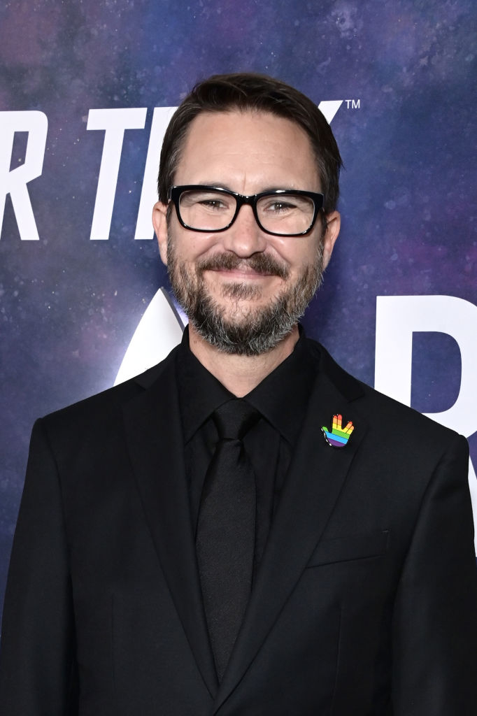 Wil Wheaton smiling on the red carpet, wearing a black suit and shirt with a colorful pin on his lapel