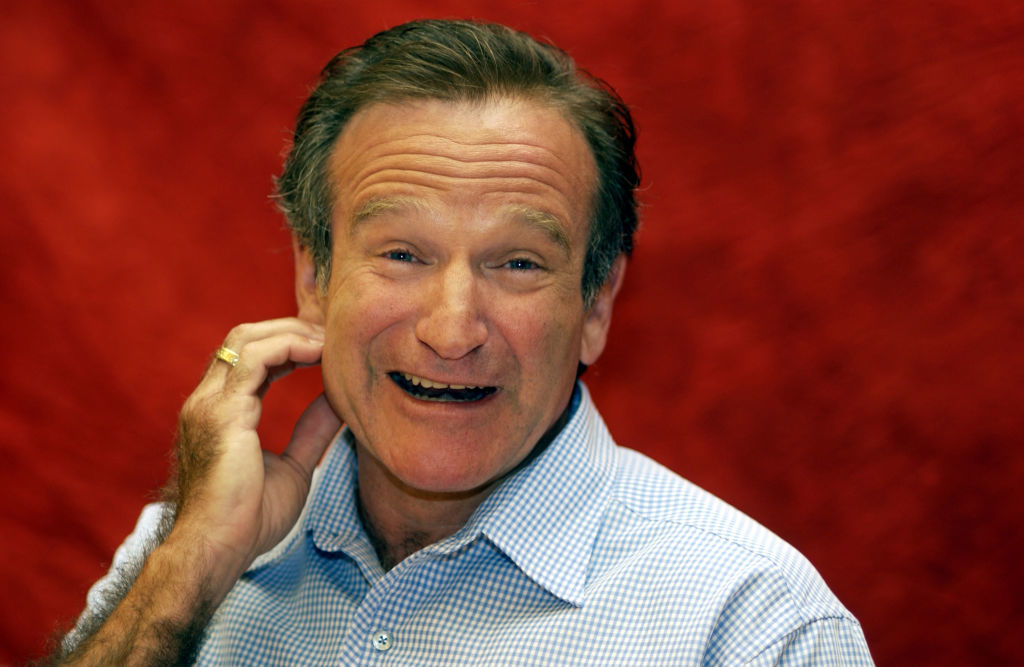 Robin Williams smiling and scratching the back of his head against a red background