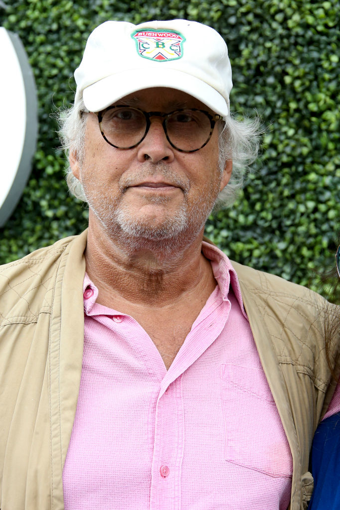 Chevy Chase in a casual outfit, wearing a white cap, glasses, a pink shirt, and a beige jacket, standing outdoors