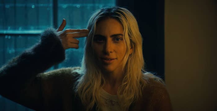 Lady Gaga pointing her fingers to her head in a gun gesture, with a serious expression, wearing a casual sweater in a scene from oker: Folie à Deux