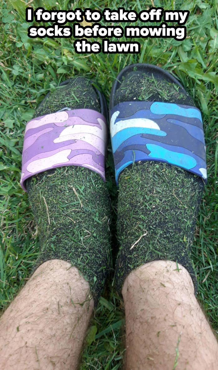 Two feet wearing mismatched slide sandals, one purple camo and one blue camo, standing on grass with grass clippings covering the socks and legs