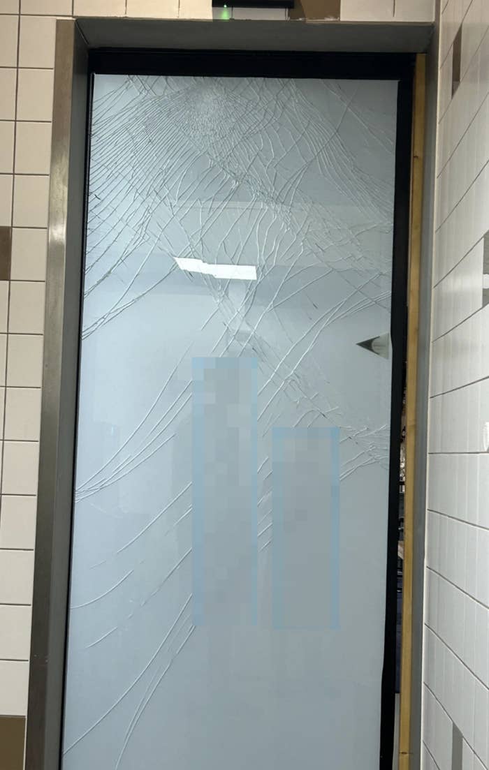 A glass door with multiple cracks and fractures is shown, with walls tiled in a square pattern surrounding the door. The glass appears severely damaged