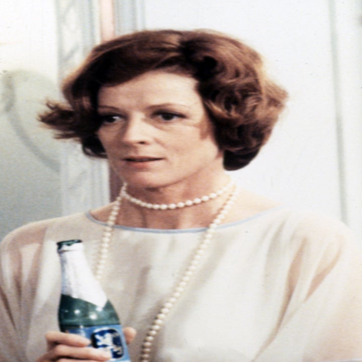 Maggie Smith holds a bottle while dressed in an elegant, sheer outfit with a pearl necklace