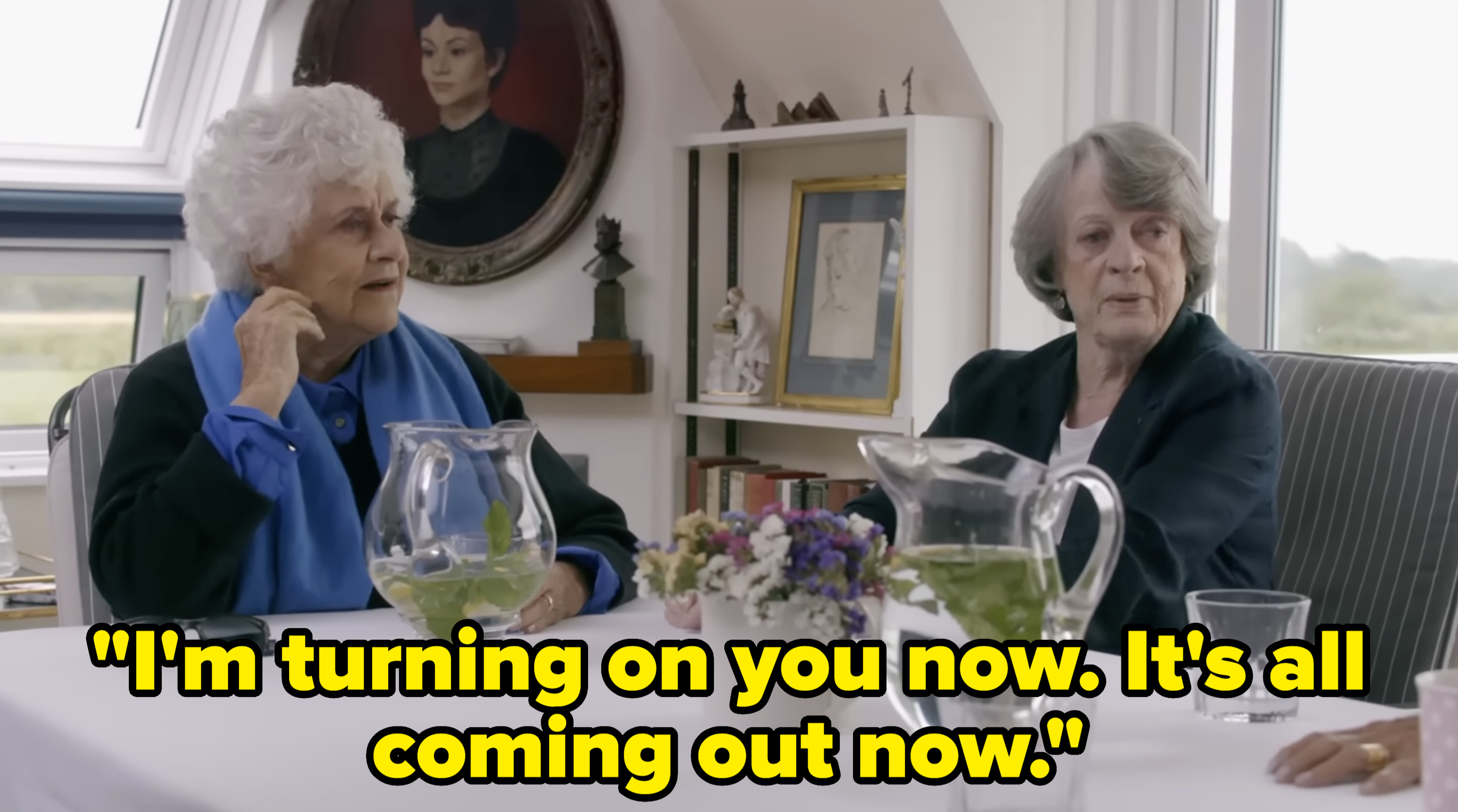 Joan and Maggie Smith sit at a table in a well-lit room, engaged in conversation. A portrait and books can be seen in the background