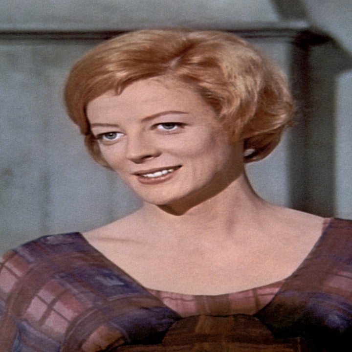 Maggie Smith in a period film, wearing a plaid dress, smiling, with two people partially visible beside her