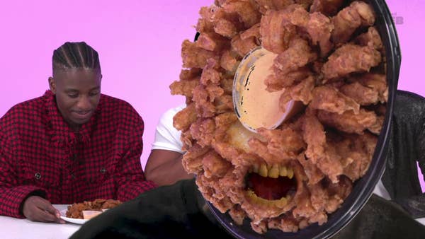 A man in a red checkered shirt sits at a table eating fried food. Another man is holding a large bloomin&#x27; onion dish in front of his face, making a playful expression