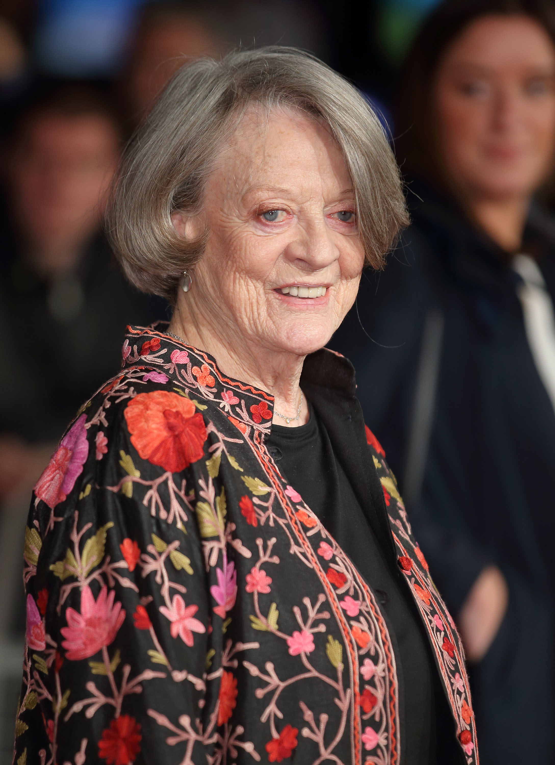 Maggie Smith smiles at the camera wearing a jacket adorned with floral embroidery at a public event