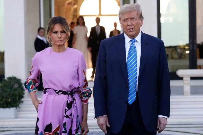 Donald Trump in a suit with a striped tie and Melania Trump in a floral dress stand outdoors in front of a building