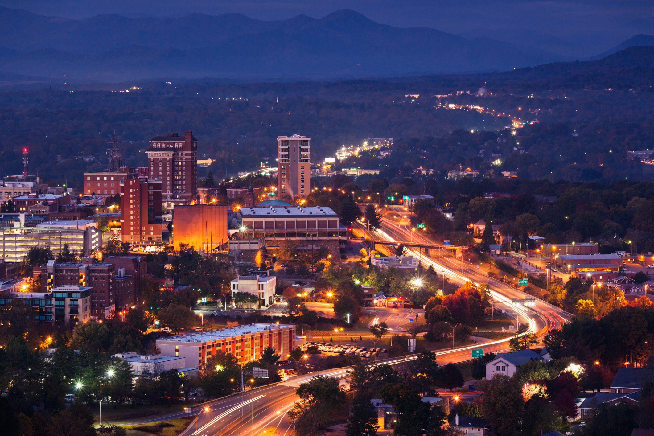 An evening cityscape of Asheville, North Carolina shows illuminated buildings, busy streets with light trails, and a mountainous background under a twilight sky