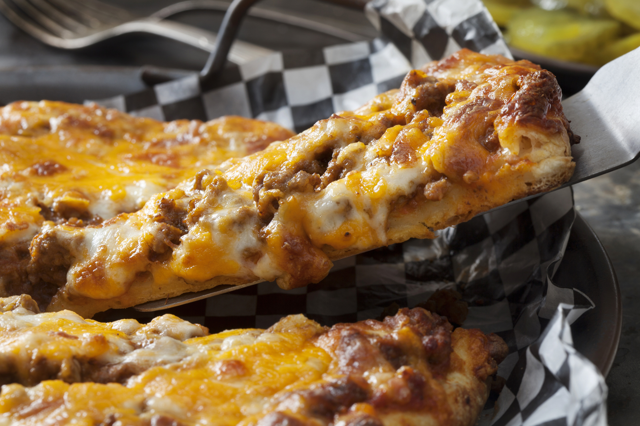 A close-up of a cheesy, meat-topped pizza slice being lifted from a checkered paper-lined plate