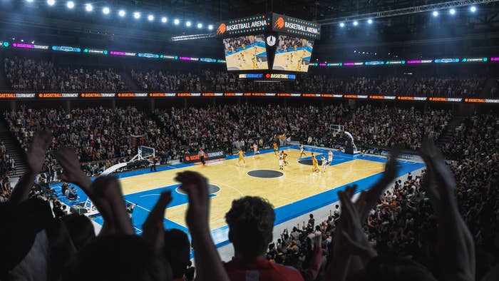 Packed basketball arena with fans cheering and a game in progress on the court. Two large screens above display the game action and the crowded stands