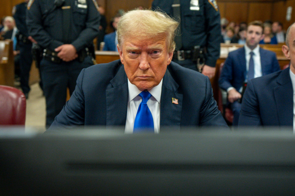 Donald Trump seated in courtroom, wearing a suit with a blue tie, focused intensely on proceedings. Police officers and other individuals visible in the background