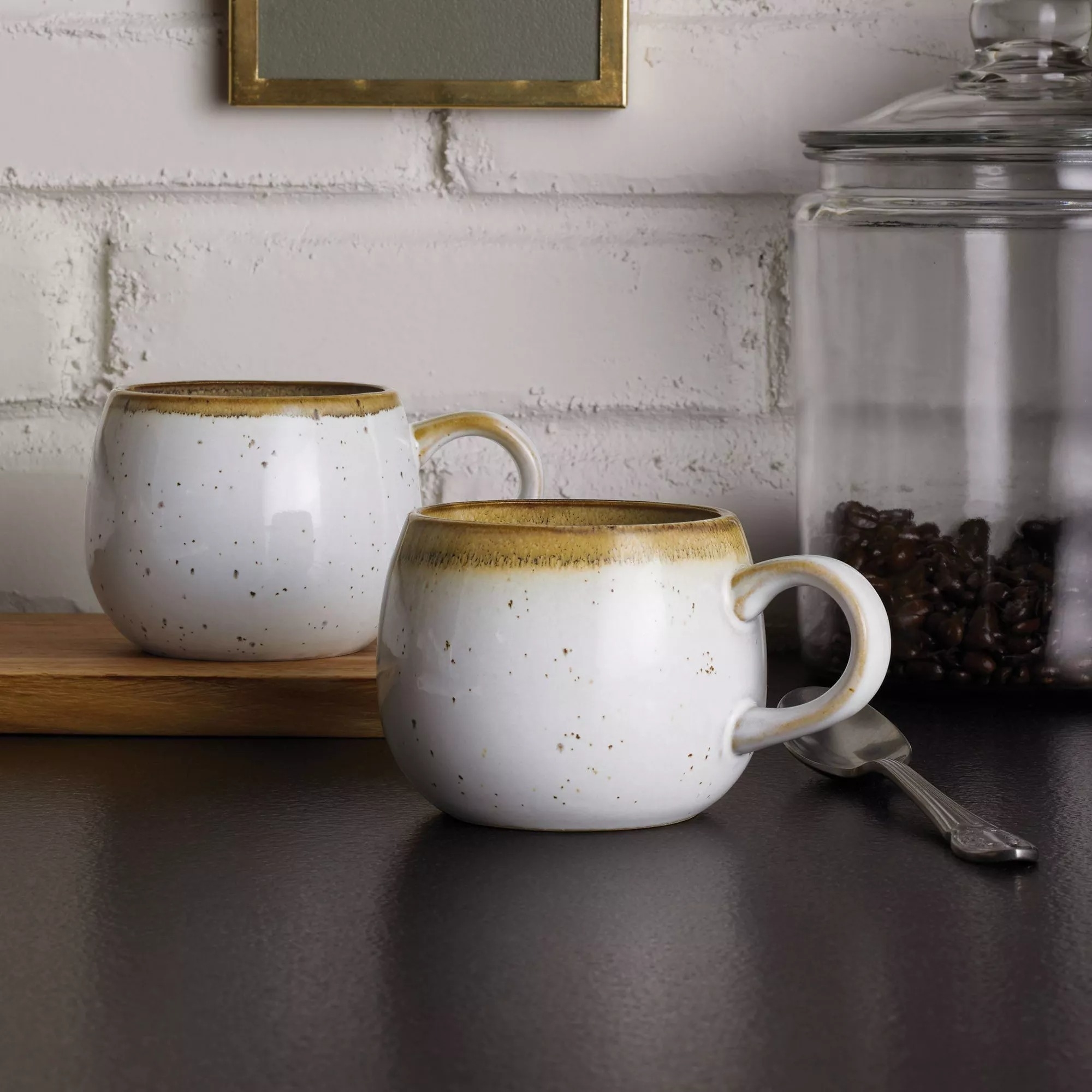 Two ceramic mugs with a rustic design are placed on a dark surface. In the background, there&#x27;s a glass jar filled with coffee beans and a wooden board