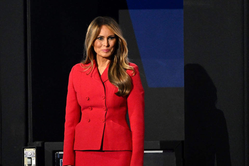 Melania Trump stands wearing a tailored, double-breasted red outfit, posing for a photo with a neutral expression