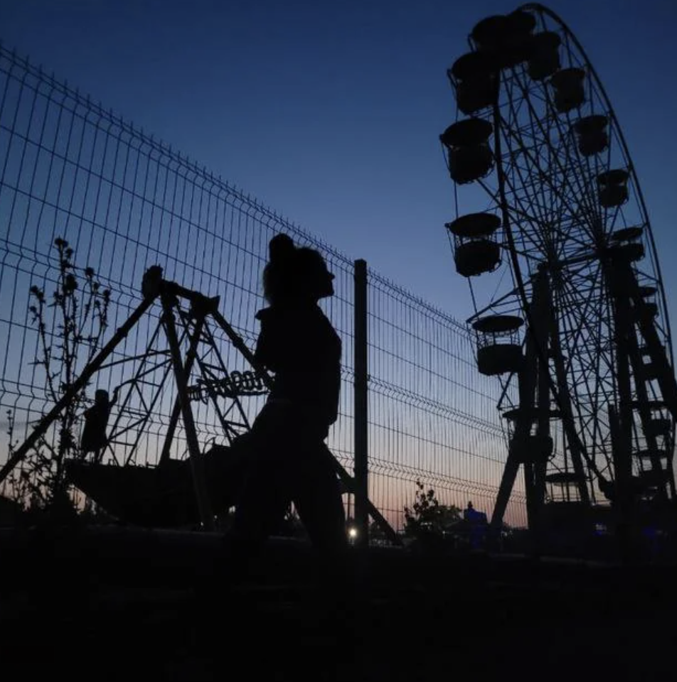 Silhouette of a woman standing near a fence with a Ferris wheel and swing ride visible at dusk in the background