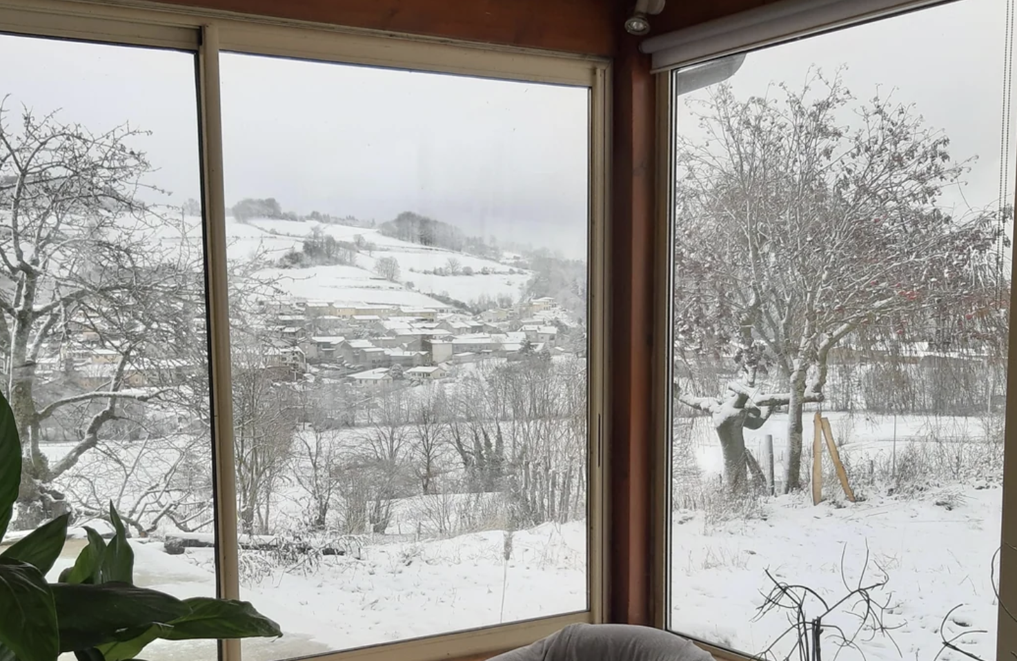 Snowy landscape viewed through large windows, showing trees, rooftops, and rolling hills covered in snow