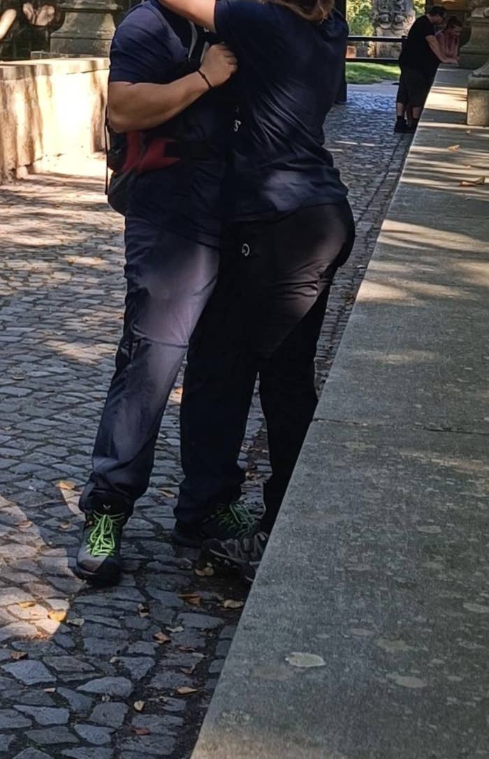 Two people, partially visible, are hugging on a cobblestone path outdoors. Both are dressed in casual clothing and dark pants
