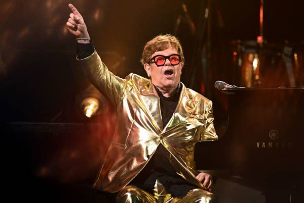 Elton John performs energetically on stage, wearing a shiny gold suit, while playing the piano