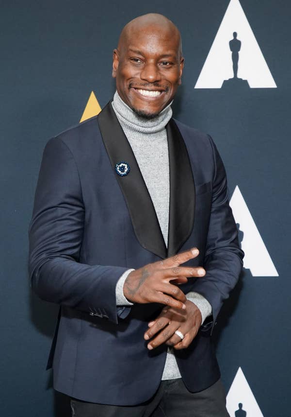 Tyrese Gibson at a formal event, smiling and posing in a dark blazer with a satin lapel over a turtleneck sweater. Oscar statue symbol in the background