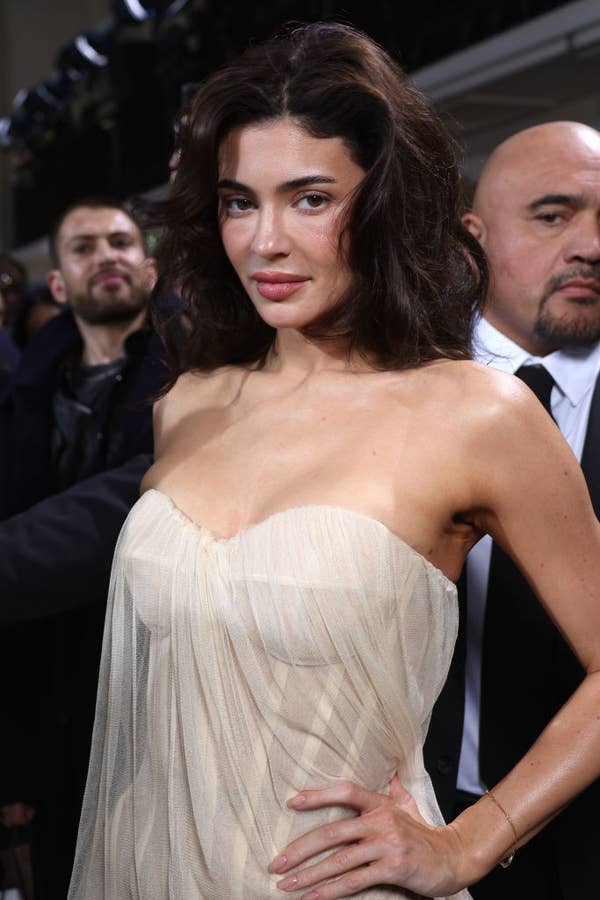A woman poses on a red carpet event in an elegant, strapless dress, surrounded by other attendees. Names are not provided