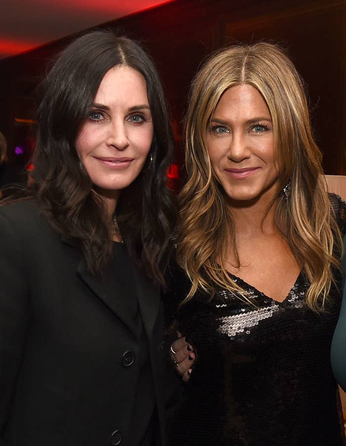 Courteney Cox and Jennifer Aniston smiling and posing at an event. Courteney wears a black blazer and Jennifer wears a black sequined dress