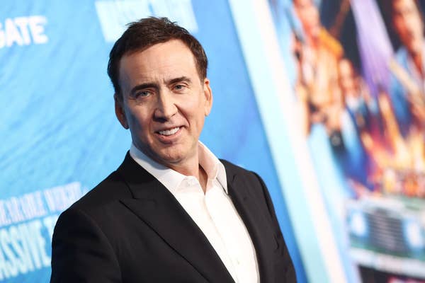Nicolas Cage at a premiere event, smiling and wearing a black suit with a white dress shirt. A blue backdrop with text and images is behind him