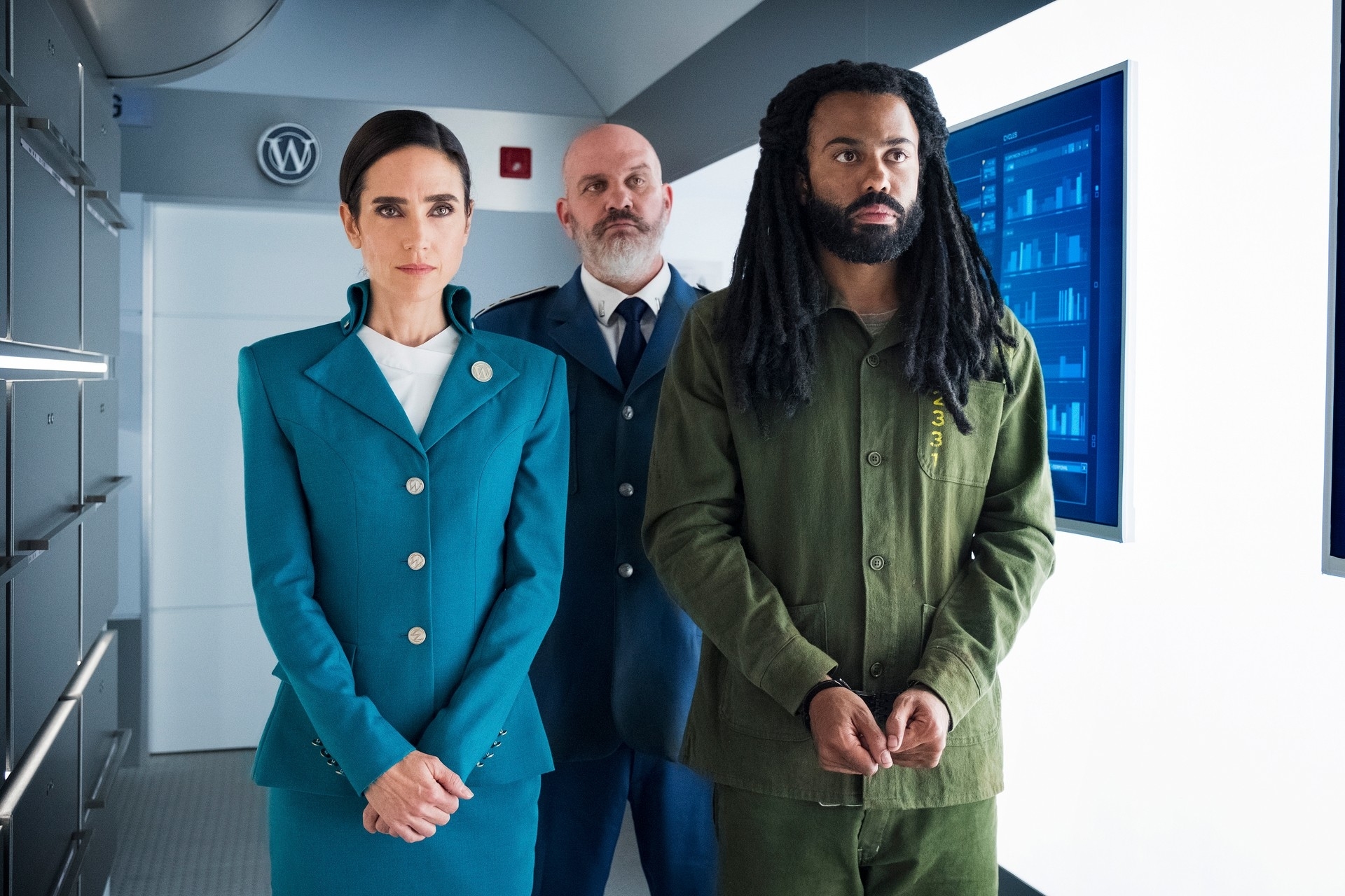 Jennifer Connelly in a teal suit, Mike O'Malley in a blue uniform, and Daveed Diggs in a green jumpsuit, standing in a futuristic setting from &quot;Snowpiercer.&quot;