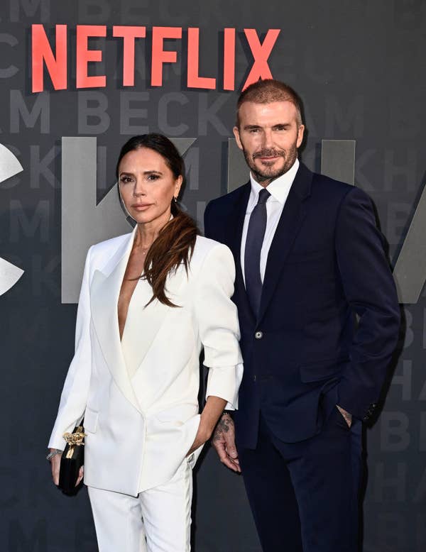 Victoria Beckham in a stylish white blazer and pants stands next to David Beckham in a dark suit at a Netflix event