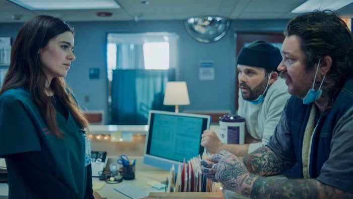 Molly Gordon in medical scrubs, speaking to a bearded man with tattoos and a mask around his neck, and another man in a beanie by a hospital desk in a scene from The Bear