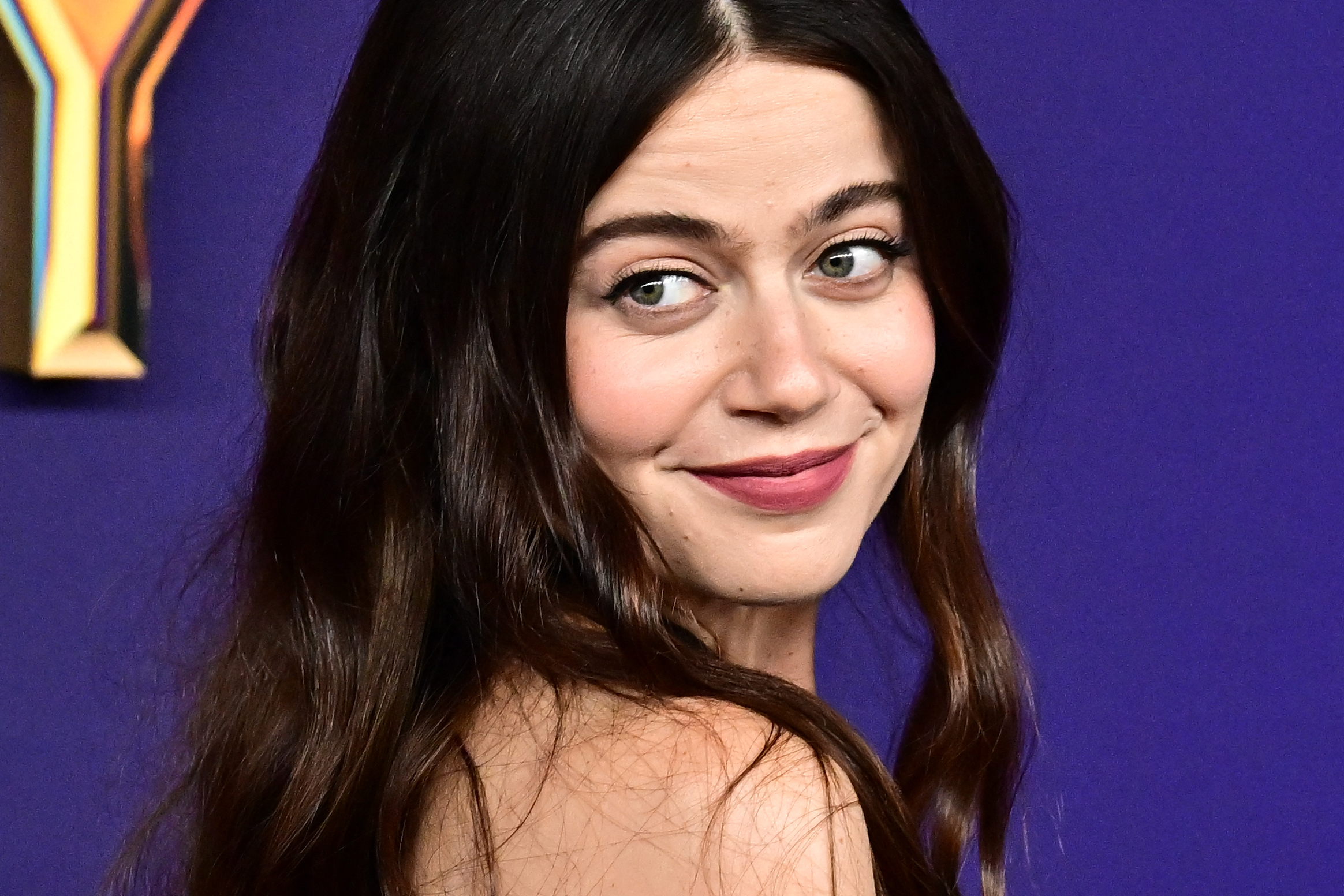 Molly Gordon poses against a background, glancing over her shoulder with a subtle smile