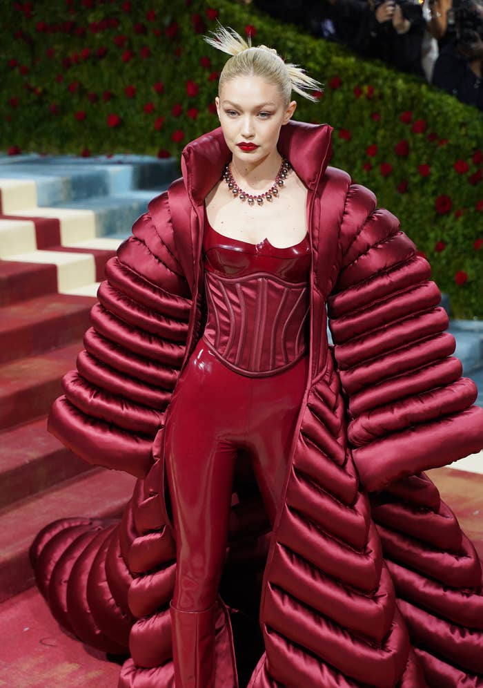 Gigi Hadid at a red carpet event in an elaborate, puffy coat and matching form-fitting outfit, accessorized with a statement necklace