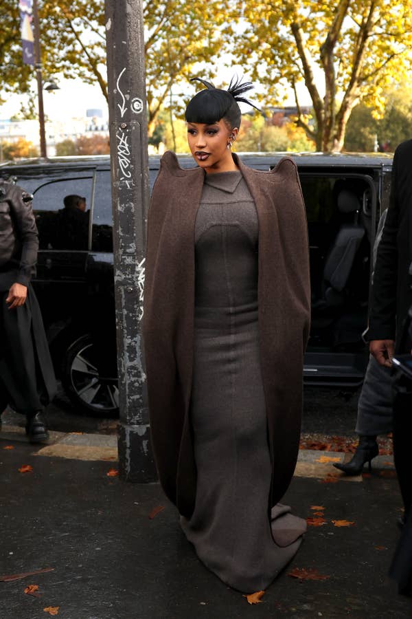 Cardi B in an avant-garde, structured outfit and headpiece, stepping out of a car, with tree-lined street in the background