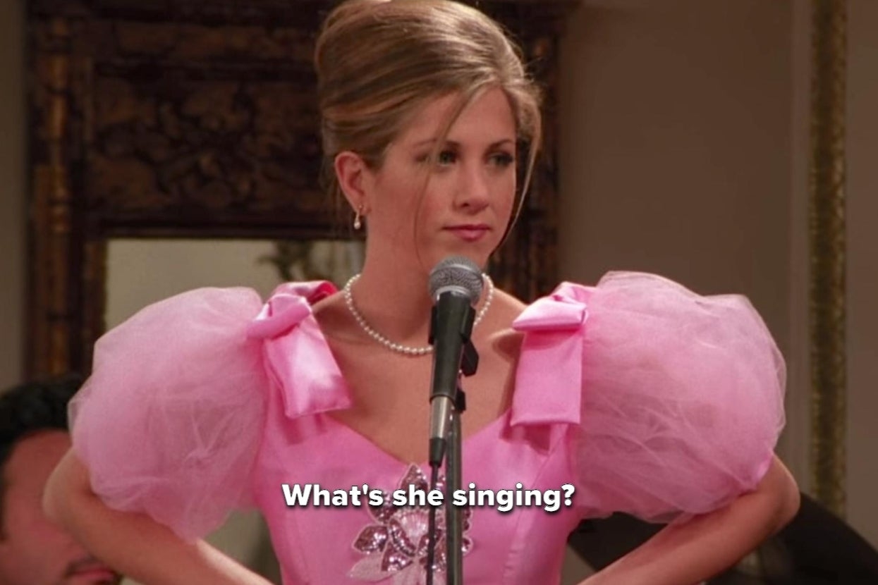 Jennifer Aniston wearing a pink dress with puffy sleeves, standing at a microphone and lip-syncing. Subtitles read: "What's she singing?"