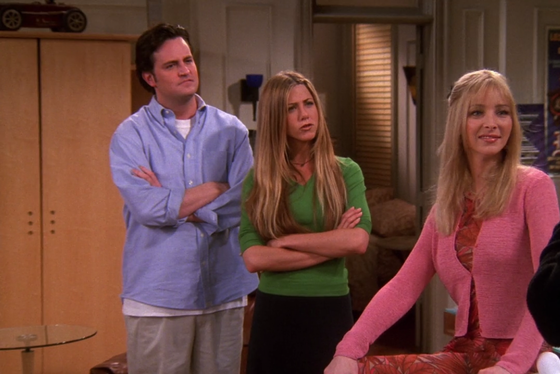 Matthew Perry, Jennifer Aniston, and Lisa Kudrow stand together in a living room, looking confused. Jennifer wears a green top, and Lisa is in a pink cardigan and floral dress